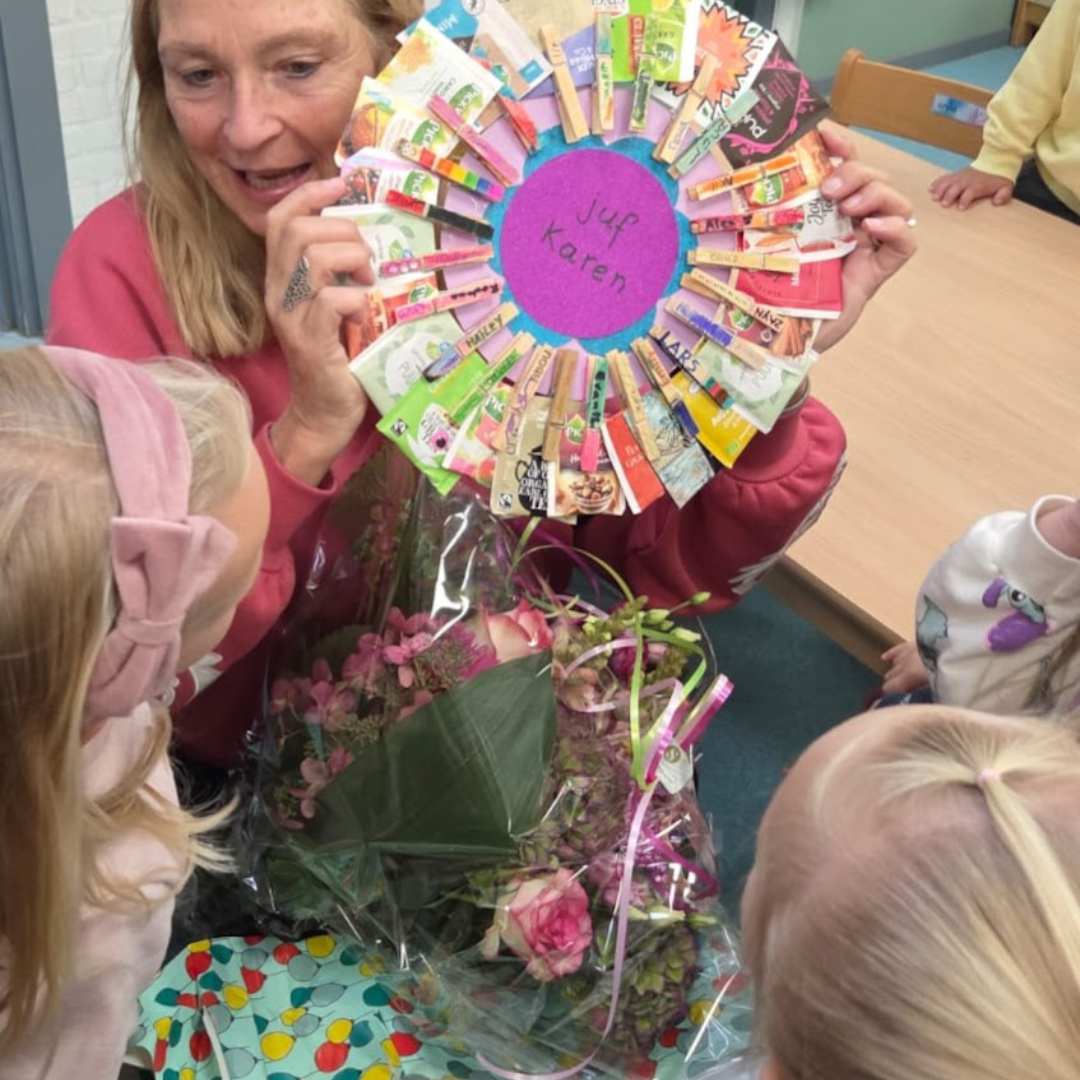 Juf Karen ontvangt bloemen en een zelfgemaakt theekrans-cadeau van haar kleutergroep als bedankje; aandacht voor veilige groepsvorming.